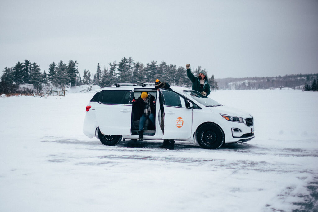 a group of people stopped on the side of the highway, winter driving in a Peg City Car Co-op van. Two of them are stepping out of the van, and the third is signalling for help.