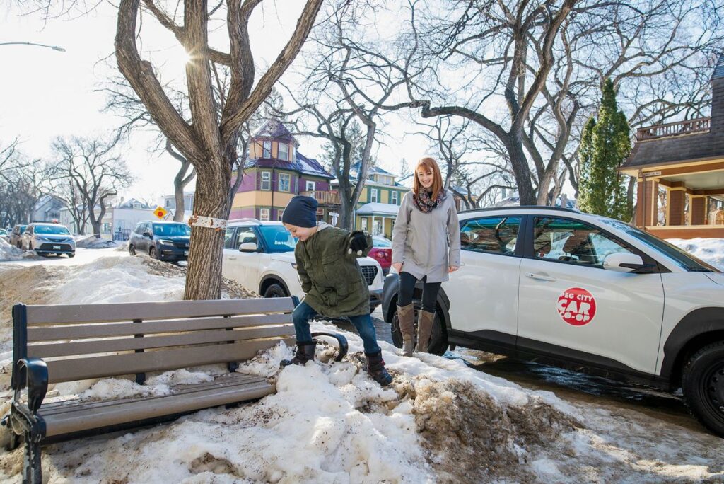 a child climbs a snow bank while a Peg City member watches over the child, standing in front of a Peg City car