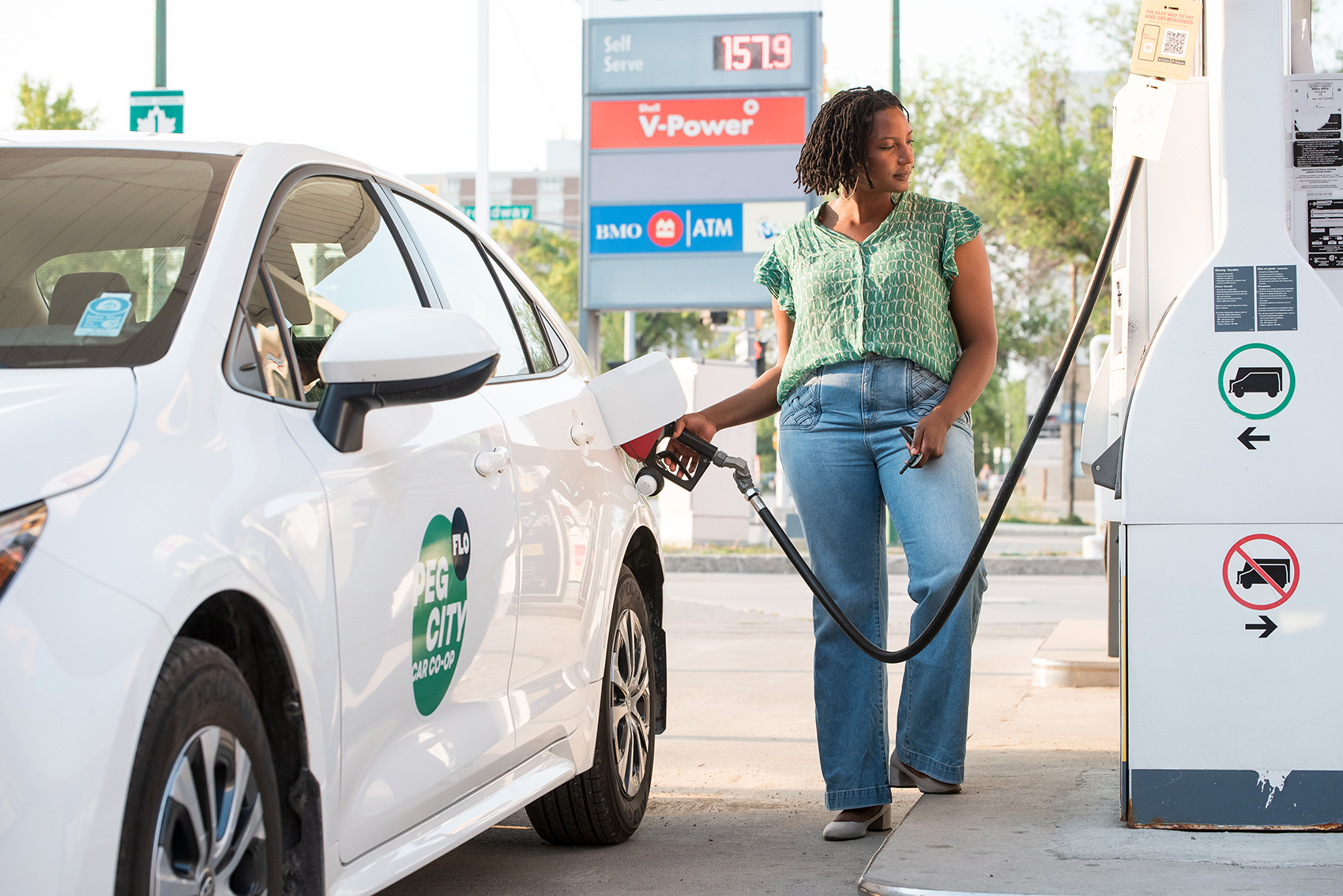 Person fueling a white car at a gas pump at a station, holding the nozzle.